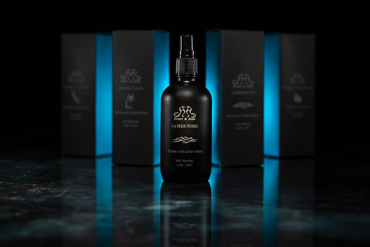 Four dark spray bottles with black pump tops arranged in a row on a glossy black reflective surface against a black background.