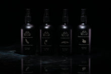 Load image into Gallery viewer, Four dark spray bottles with black pump tops arranged in a row on a glossy black reflective surface against a black background. Each bottle has a white printed logo and text, including names such as  "Santa Coco,” “La Mer Noire,” and “Cérdrat et Mandarine" and Crème de Lavande".