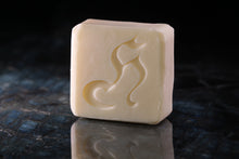 Load image into Gallery viewer, A rectangular bar of cream-colored soap with an embossed stylized design, standing on a dark reflective surface against a dark background.