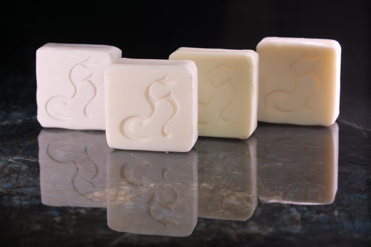 Four square bars of soap in off-white and cream shades, each embossed with a stylized cat silhouette, arranged on a dark reflective surface against a black background.