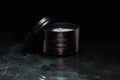 A black metal tin candle with its lid leaning against it, set on a dark reflective surface against a black background. The candle has white wax and gold printed text and logo reading 'La Mer Noire'.