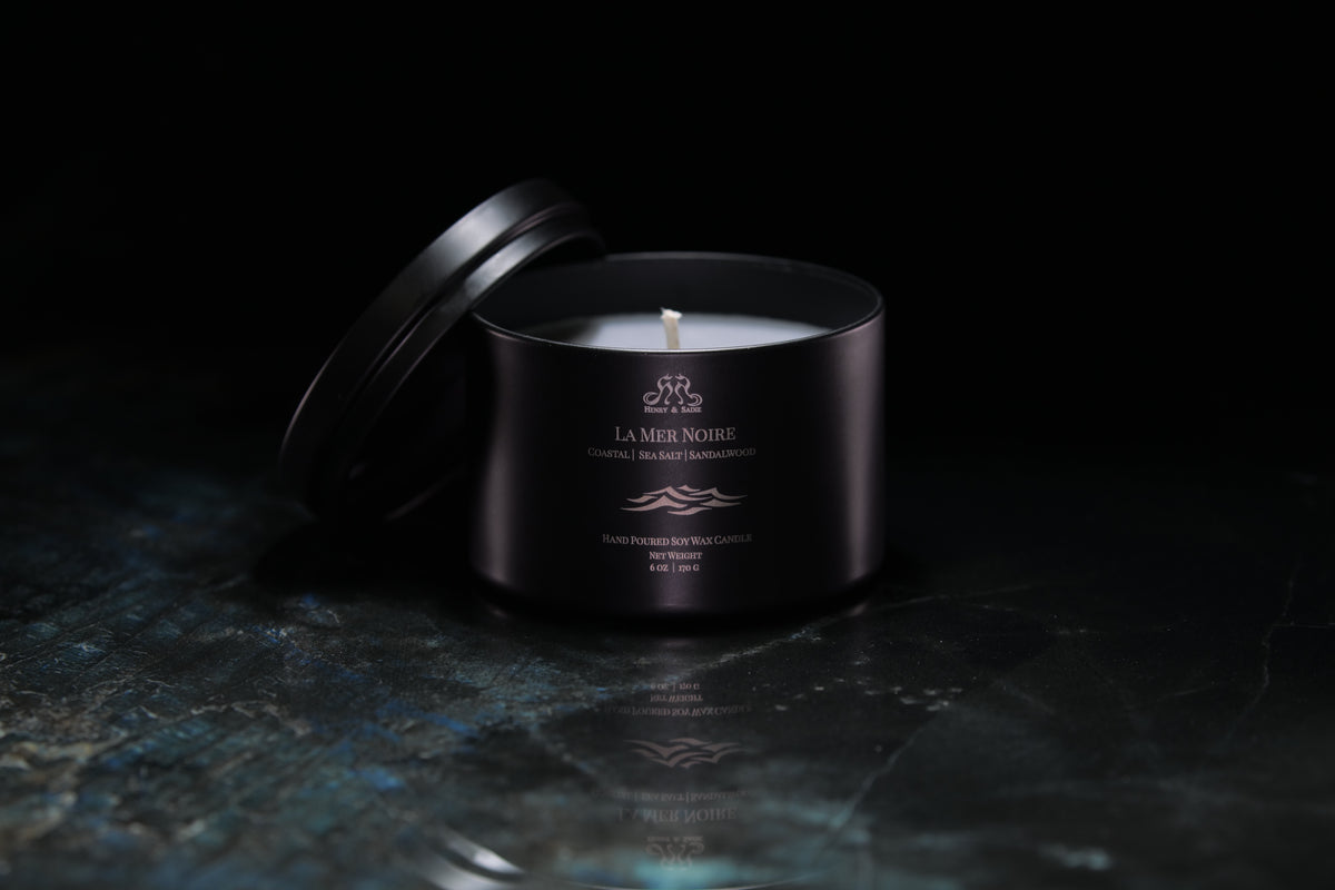 A black metal tin candle with its lid leaning against it, set on a dark reflective surface against a black background. The candle has white wax and gold printed text and logo reading 'La Mer Noire'.