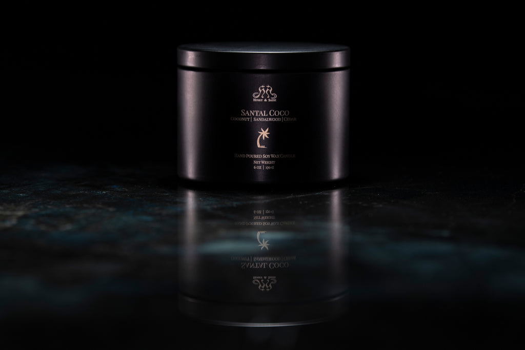 A black metal candle tin with its lid closed. The tin is labeled 'SANTAL COCO' and sits on a dark, reflective marble-like surface against a black background.