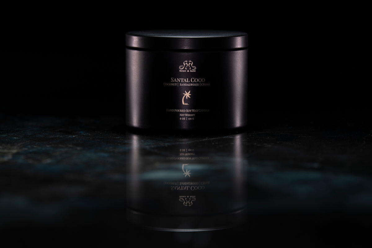 A black metal candle tin with its lid closed. The tin is labeled 'SANTAL COCO' and sits on a dark, reflective marble-like surface against a black background.