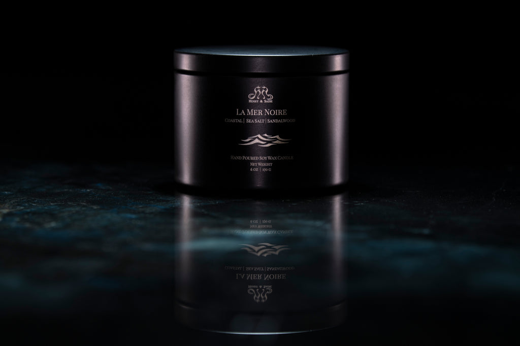 A black metal tin candle with its lid on, set on a dark reflective surface against a black background. The candle has white wax and gold printed text and logo reading 'La Mer Noire'.