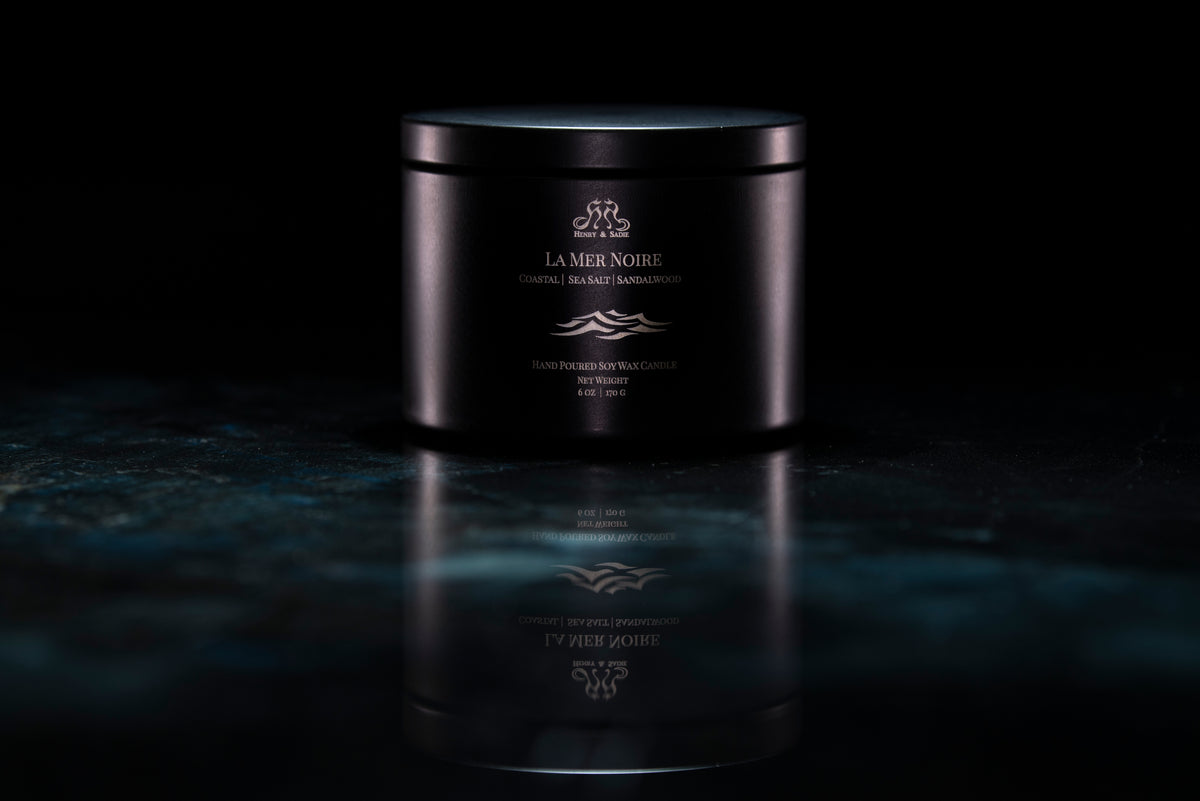 A black metal tin candle with its lid on, set on a dark reflective surface against a black background. The candle has white wax and gold printed text and logo reading 'La Mer Noire'.