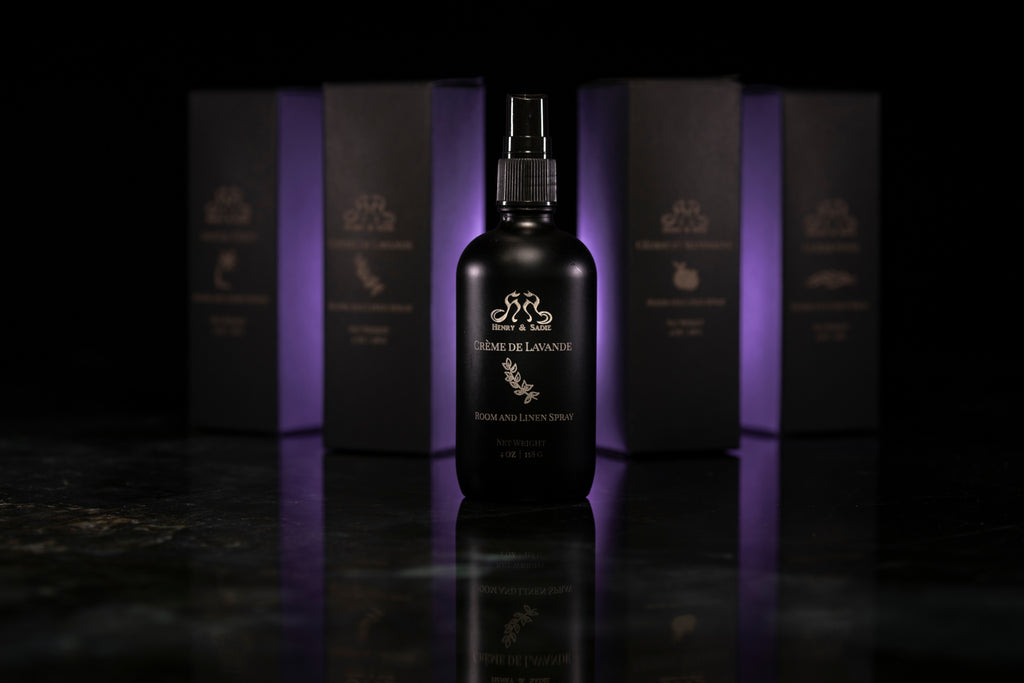 A dark bottle of Henry & Sadie Crème de Lavande Room and Linen Spray stands centered in front of several matching dark boxes with purple lighting, on a glossy black reflective surface.