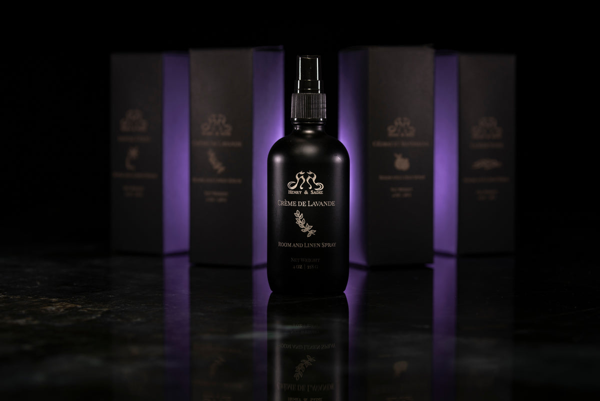 A dark bottle of Henry & Sadie Crème de Lavande Room and Linen Spray stands centered in front of several matching dark boxes with purple lighting, on a glossy black reflective surface.