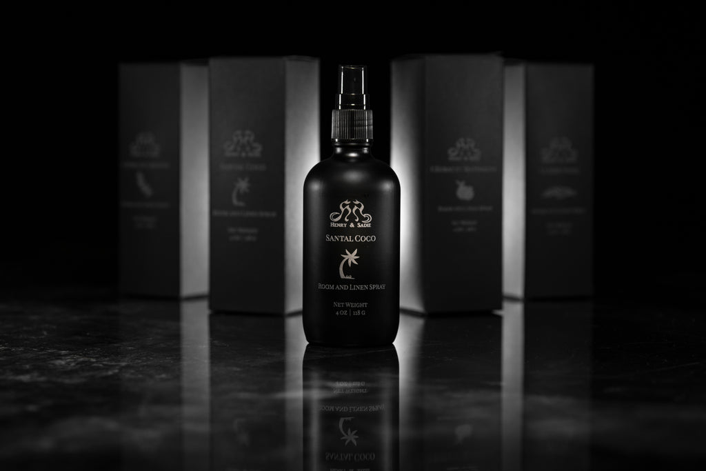 A matte black spray bottle labeled Santal Coco Room and Linen Spray stands centered on a reflective dark surface, with several matching black product boxes blurred in the background.