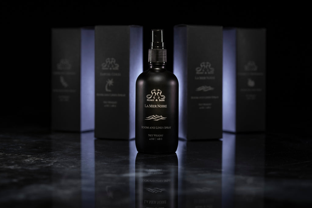A black bottle of Henry & Sadie La Mer Noire Room and Linen Spray sitting on a black and white reflective surface with similar boxes blurred out in the background
