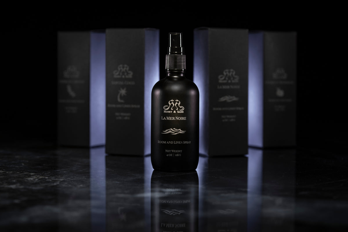 A black bottle of Henry & Sadie La Mer Noire Room and Linen Spray sitting on a black and white reflective surface with similar boxes blurred out in the background