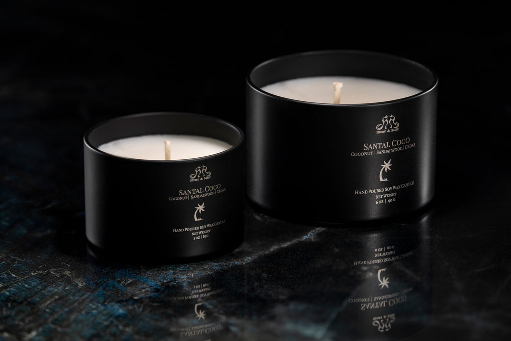 Two black tin candles with white wax and gold Santal Coco branding, displayed on a dark reflective surface against a black background.