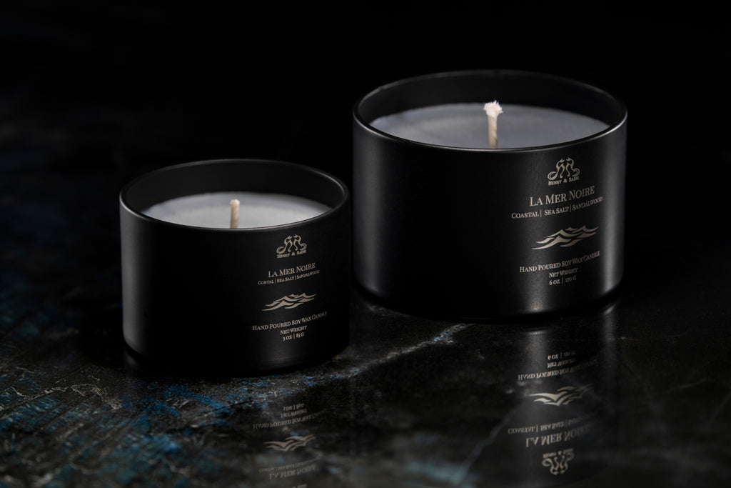 Two matte black La Mer Noire candles on a dark reflective surface against a black background.
