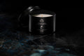 A black metal candle tin with its lid propped open, showing a white wax candle inside. The tin is labeled 'SANTAL COCO' and sits on a dark, reflective marble-like surface against a black background.