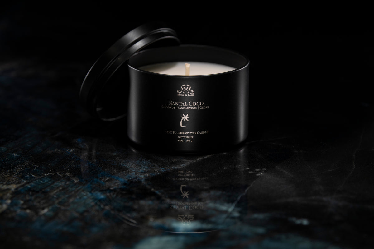 A black metal candle tin with its lid propped open, showing a white wax candle inside. The tin is labeled 'SANTAL COCO' and sits on a dark, reflective marble-like surface against a black background.