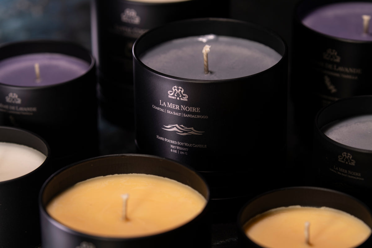 An assortment of candles with gray, pale yellow, lavender, and white wax arranged with the "La Mer Noire" candle in the center in a black tin
