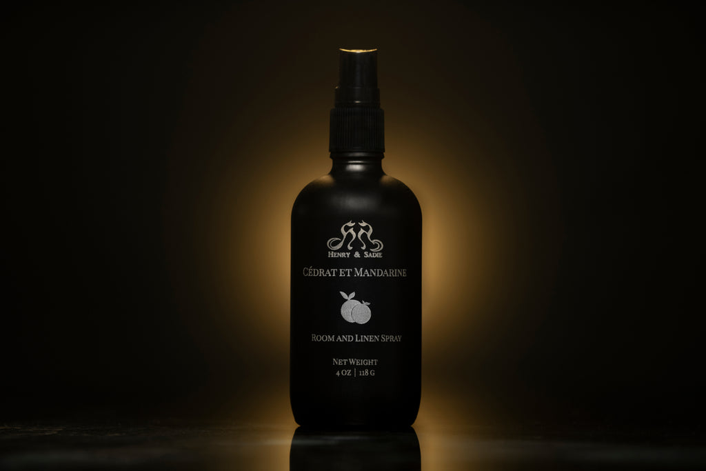 A black room and linen spray bottle standing against a dark background with a warm spotlight glow behind it. The bottle features white text and a small white mandarin fruit illustration, with the Henry & Sadie brand 'Cédrat et Mandarine' visible.