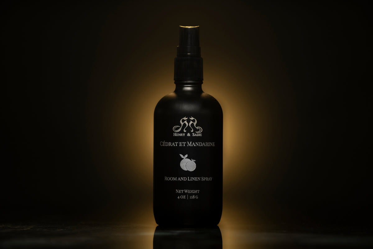 A black room and linen spray bottle standing against a dark background with a warm spotlight glow behind it. The bottle features white text and a small white mandarin fruit illustration, with the Henry & Sadie brand 'Cédrat et Mandarine' visible.