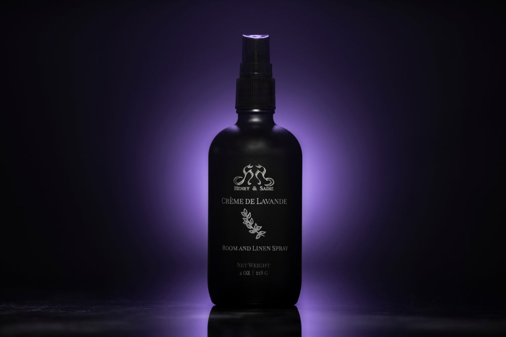 A black spray bottle of Henry & Sadie Crème de Lavande room and linen spray, illuminated by a purple glow against a dark background.