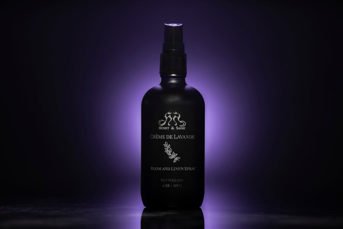 A black spray bottle of Henry & Sadie Crème de Lavande room and linen spray, illuminated by a purple glow against a dark background.
