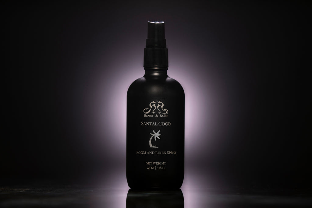 A matte black spray bottle of Santal Coco Room and Linen Spray, centered against a dark background with a soft  glow behind it.