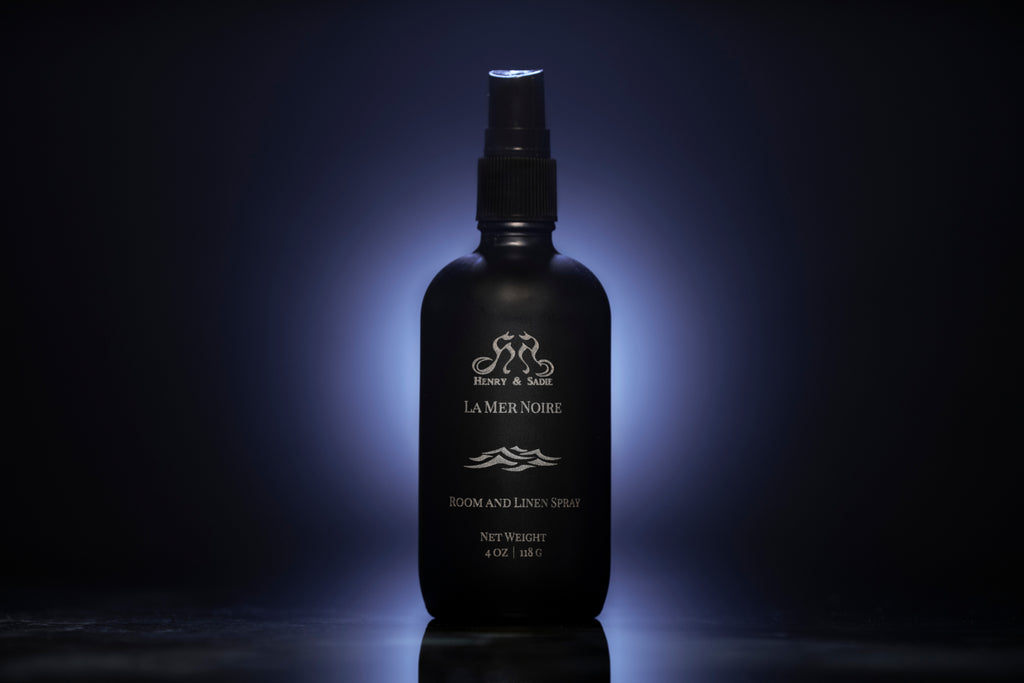 A black bottle of La Mer Noire Henry & Sadie Room Spray on a black reflective surface with a soft blue gray glow in in the background