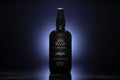 A black bottle of La Mer Noire Henry & Sadie Room Spray on a black reflective surface with a soft blue gray glow in in the background