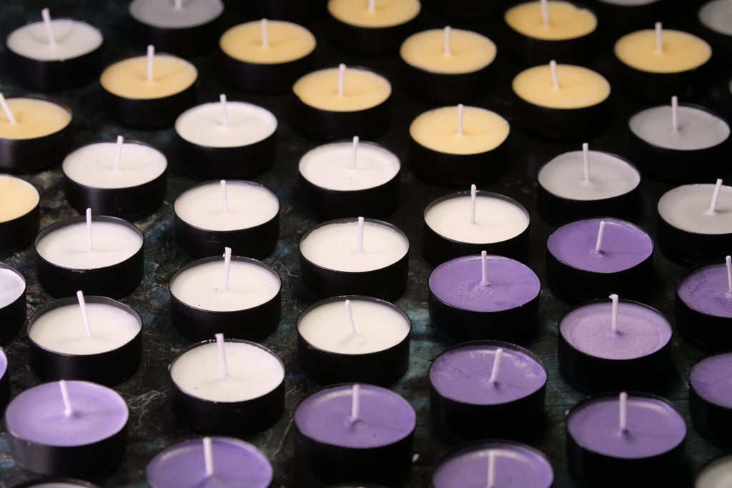 A close-up arrangement of small tealight candles in black holders, shown in white, lavender purple, and pale yellow tones on a dark surface.