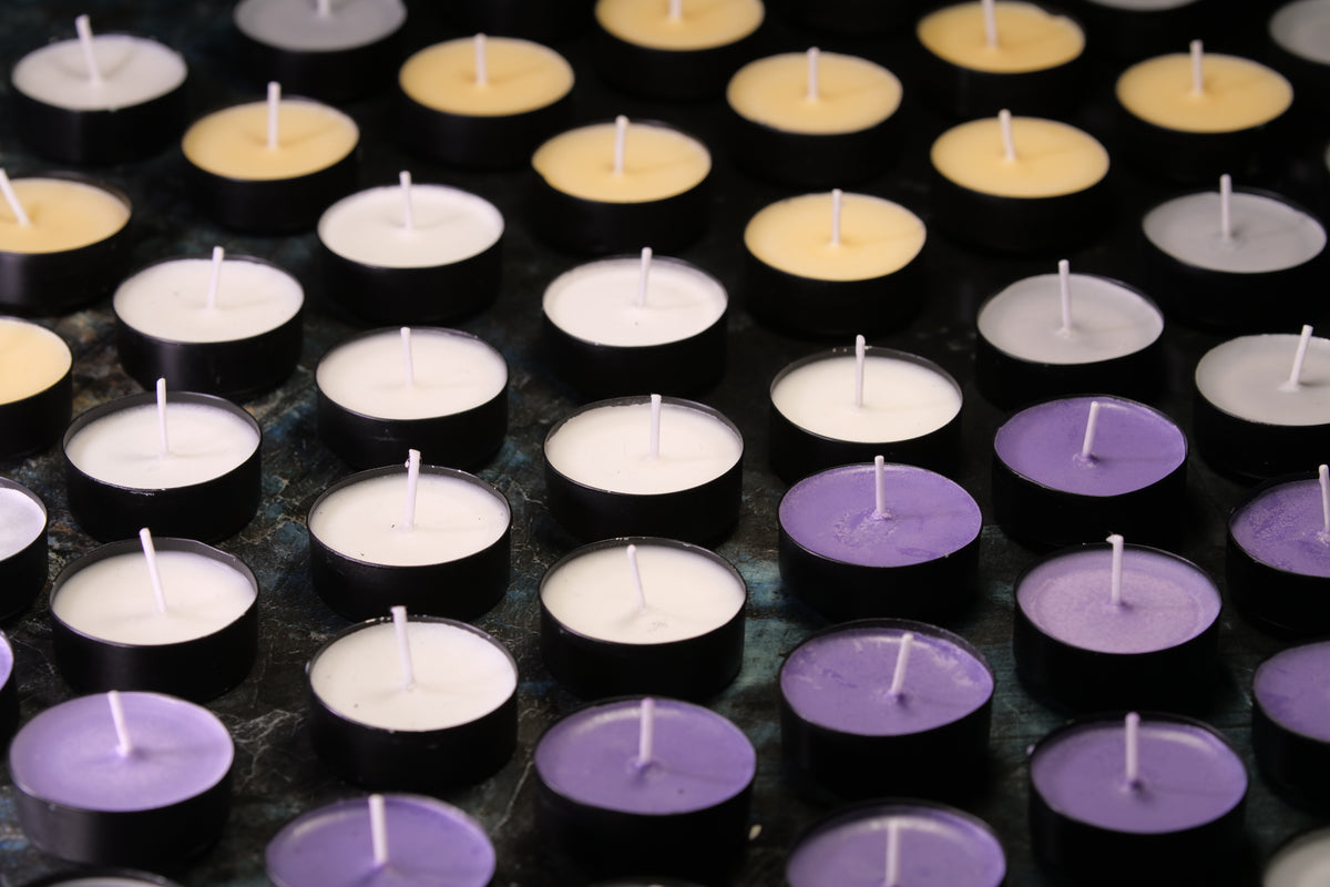 A close-up arrangement of small tealight candles in black holders, shown in white, lavender purple, and pale yellow tones on a dark surface.