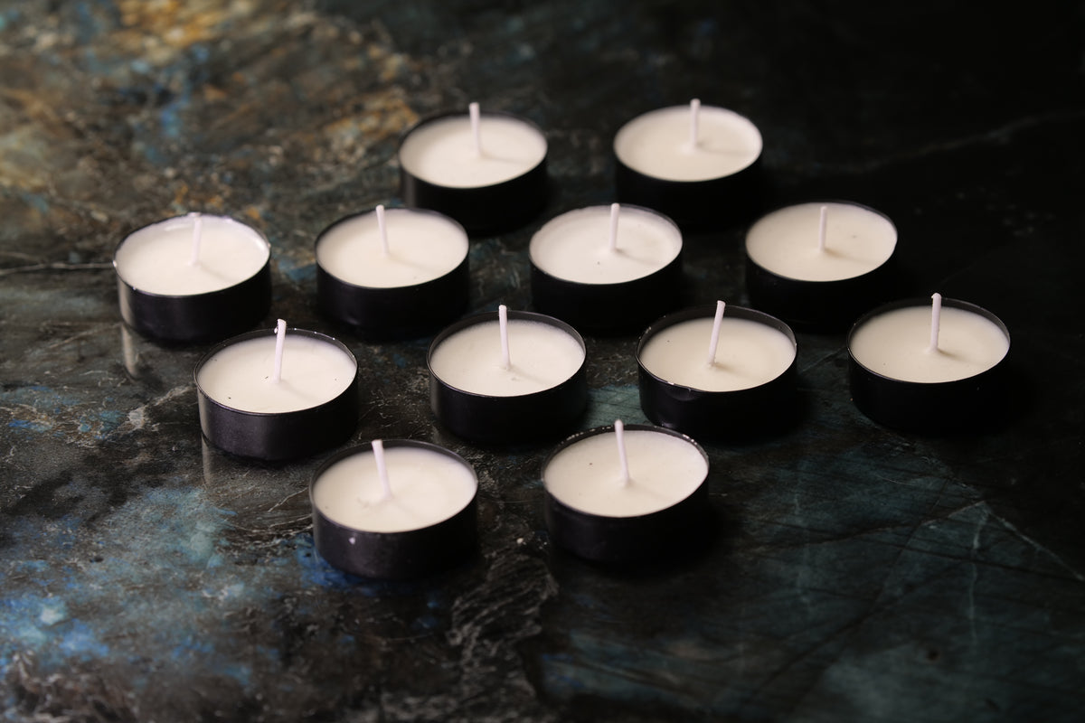 Twelve small black tea light candles with white wax are arranged on a dark, textured blue and black surface.