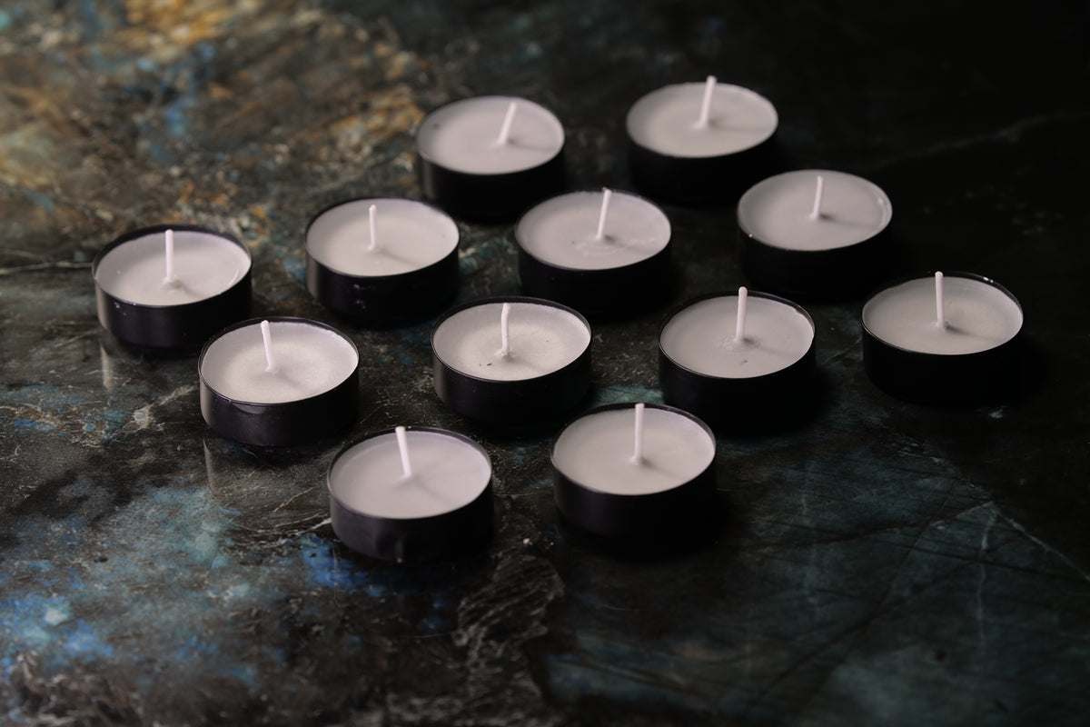 Twelve black tea light candles with light gray wax arranged on a dark, textured blue and black surface.