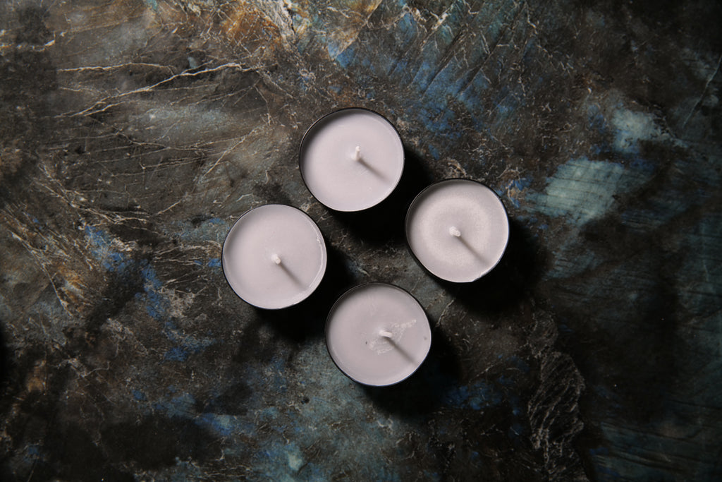 Four small tealight candles with pale gray wax and black metal holders, arranged in a loose square on a dark textured surface with blue and gray marbling.