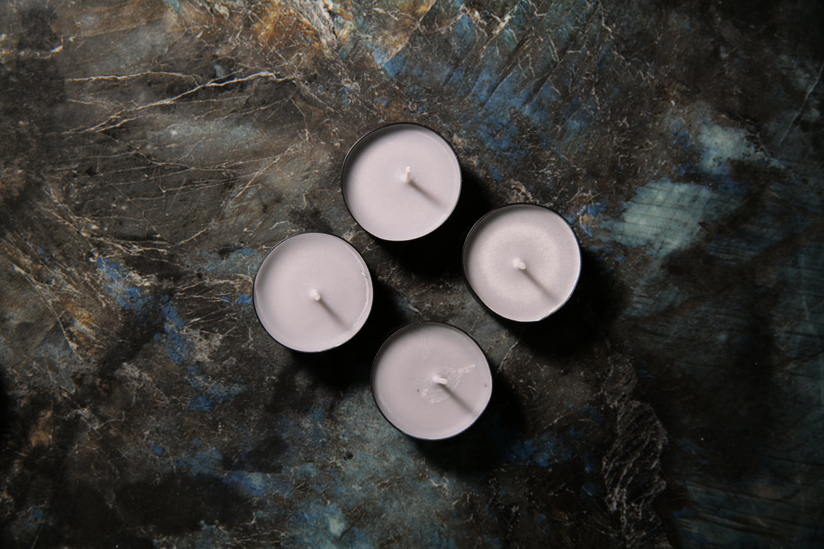 Four small tealight candles with pale gray wax and black metal holders, arranged in a loose square on a dark textured surface with blue and gray marbling.