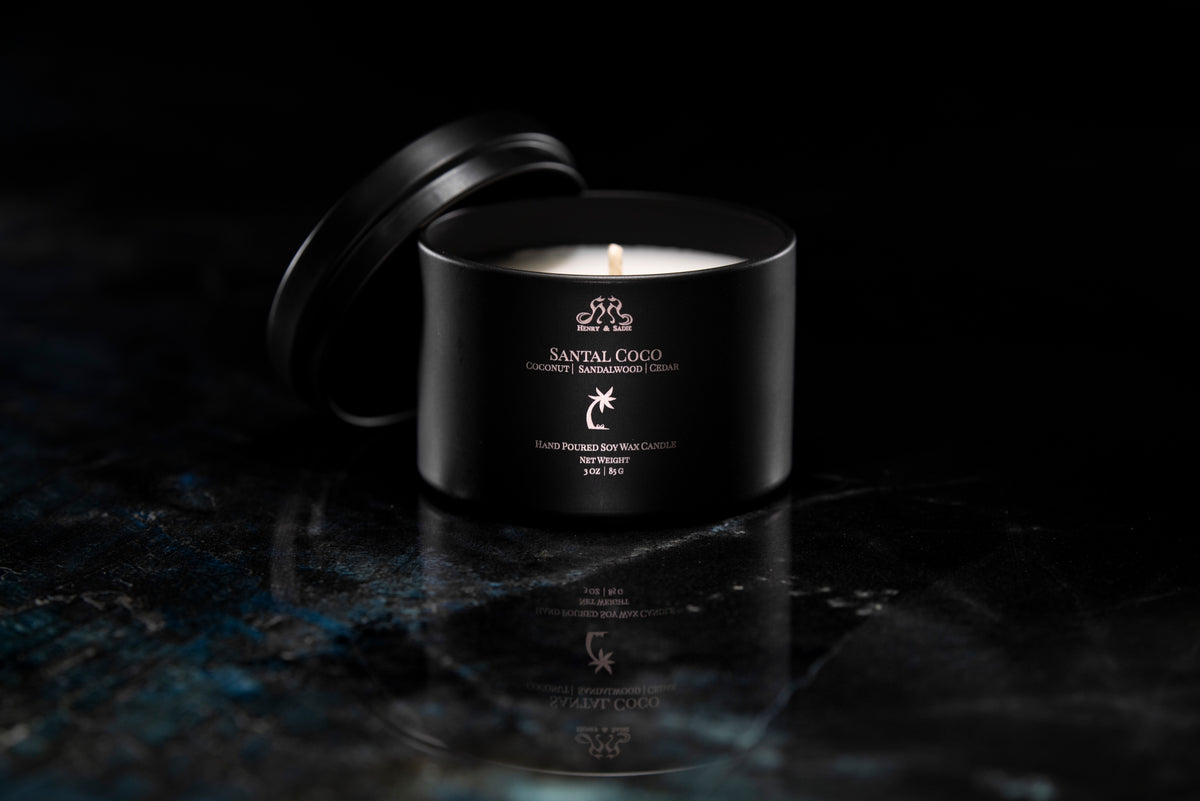 A black metal candle tin with its lid off, showing a lit white candle inside. The tin is labeled 'SANTAL COCO' and sits on a dark reflective surface against a black background.
