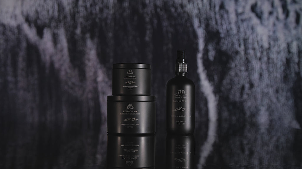 Video showing the Henry & Sadie La Mer Noire Room Spray and Candles in black containers on a reflective black surface with white and black waves crashing in the background
